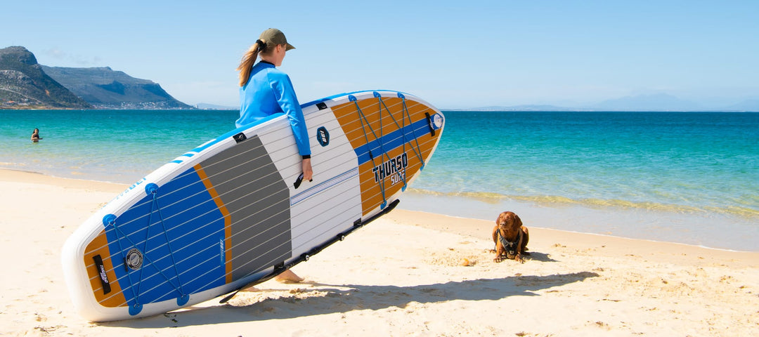 Woman approaching a dog near the shoreline while carrying a big paddleboard