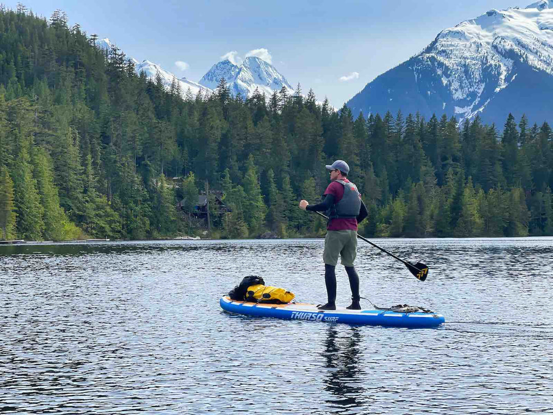 Paddleboarder on a Thurso Surf Max multi-purpose SUP paddles with his overnight camping trip gear on deck with mountains in the background