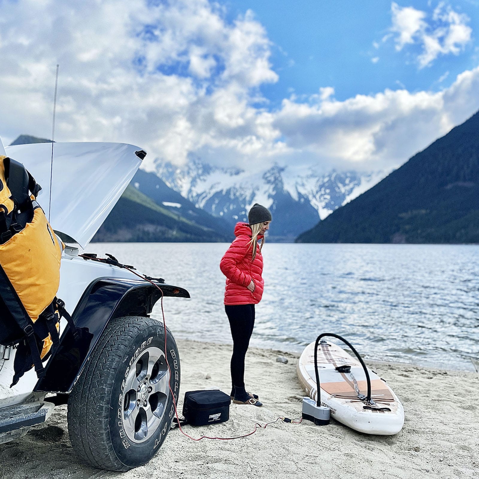 Woman standing in front of jeep on the beach with mountain and lake in the background while she waits for her Thurso Surf Waterwalker all-around SUP to inflate using an electric pump