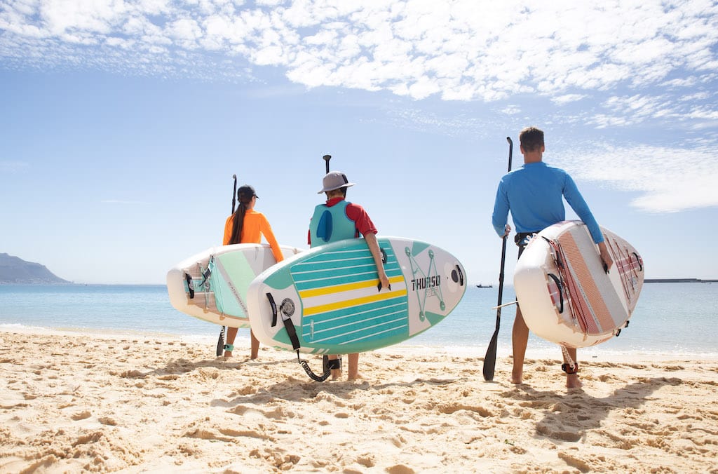 Three people holding Thurso Surf stand up paddleboards on shore while looking out to the water