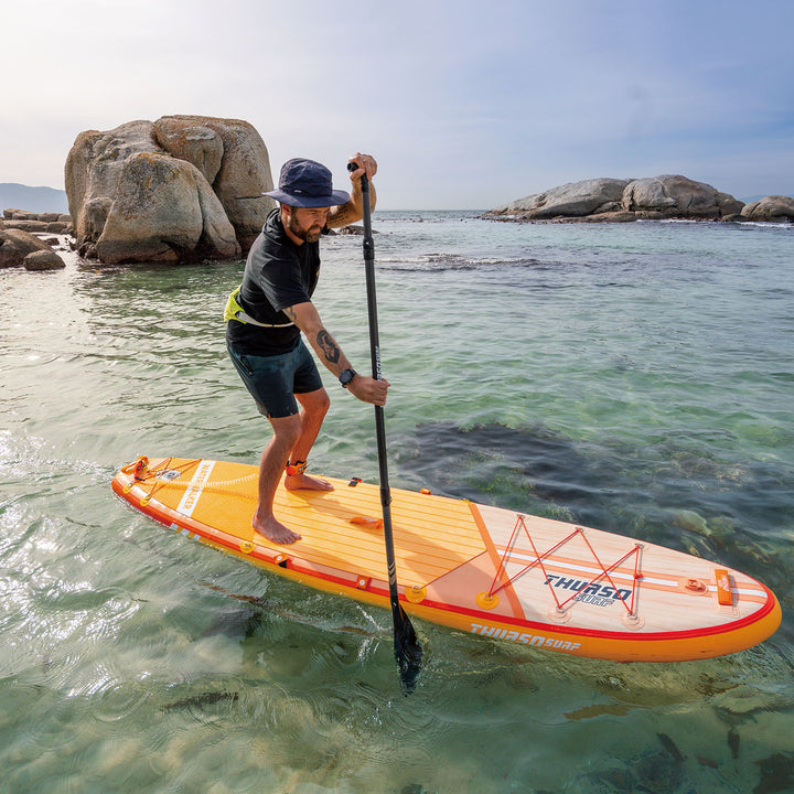 Man on a Thurso Sports tangerine paddleboard in clear water with rocks in the background