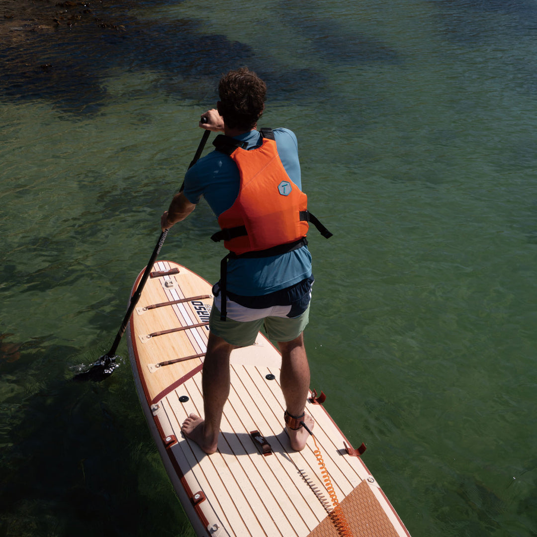 Person paddleboarding on a calm body of water wearing an orange life jacket with crimson sup.