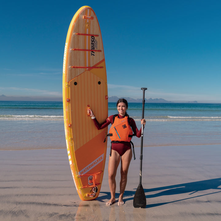 Woman holding a paddle and a large tangerine paddleboard on a beach with clear blue sky.