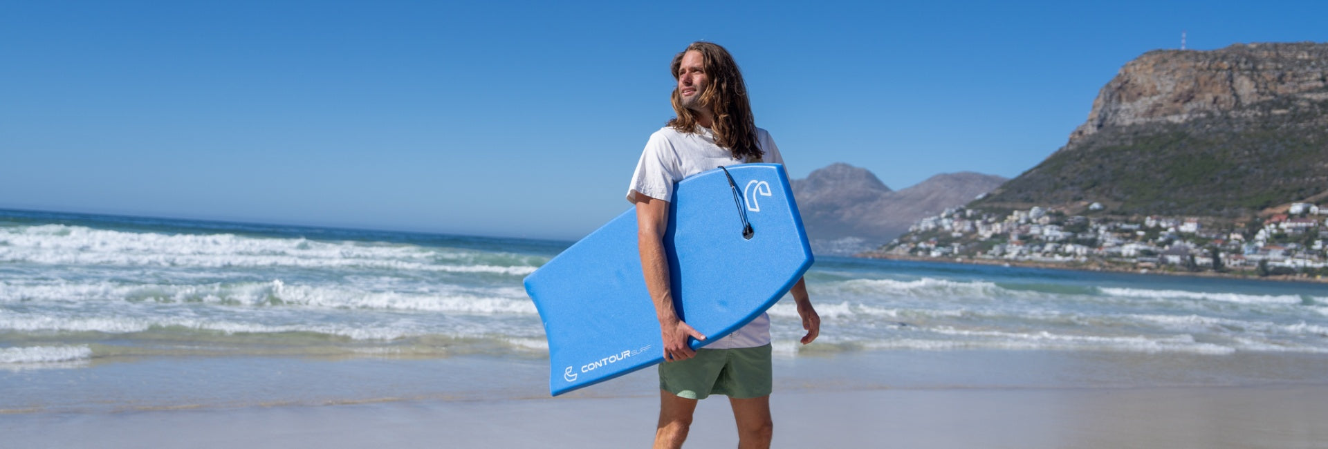 Person holding a blue bodyboard on a beach with ocean and mountains in the background