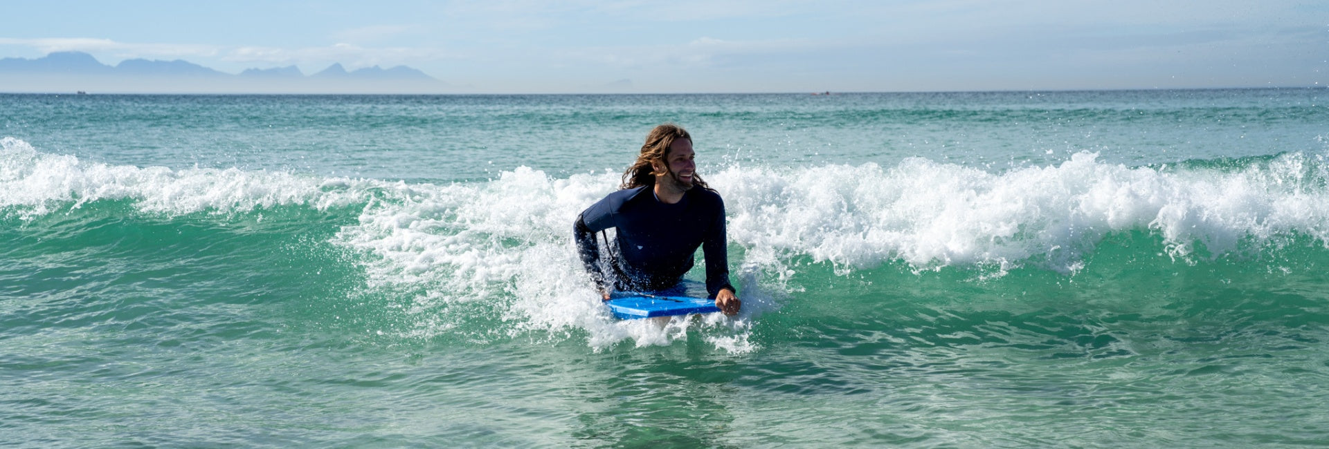 Person in a wetsuit on a bodyboard in the ocean with waves and clear sky.