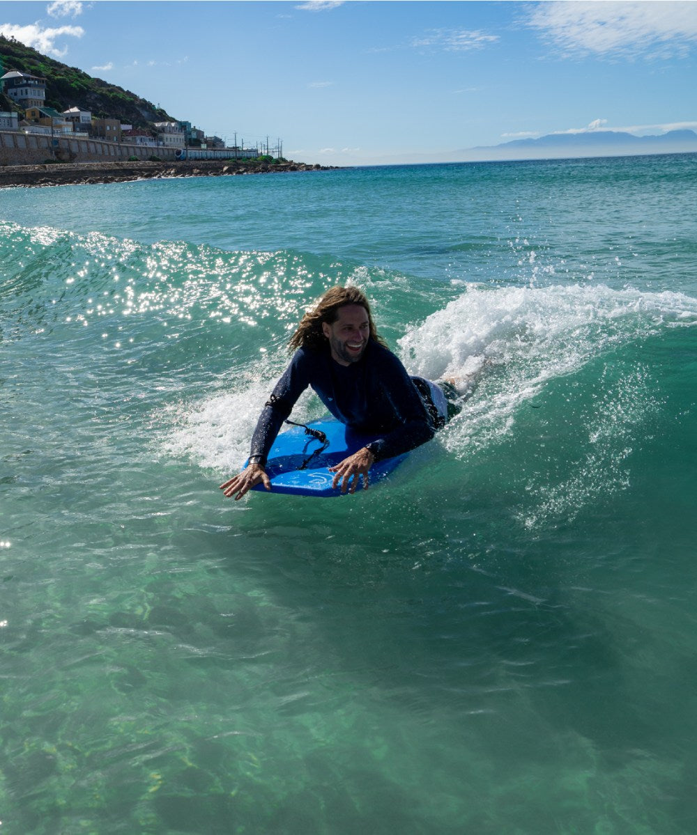 Person on a blue bodyboard riding a wave in clear water with a coastal background