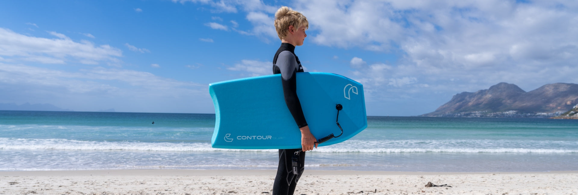 Person holding a blue bodyboard on a beach with mountains in the background