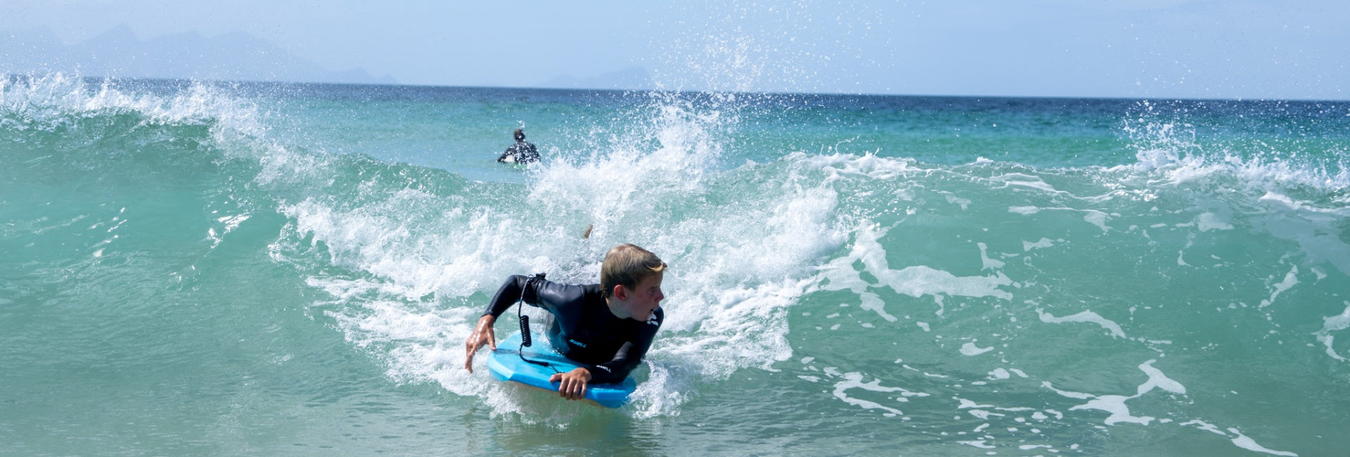 Person surfing on a blue bodyboard in the ocean with another surfer in the distance.